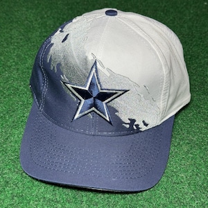 Op de afbeelding: Een marineblauwe en witte baseballcap met een geborduurd sterlogo. De cap heeft een gebogen klep en een knoop aan de bovenkant. Het ontwerp bevat een kaartachtig patroon op het witte deel.