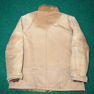 Może przedstawiać: Beżowa kurtka robocza z brązowym kołnierzem i mankietami. Kurtka nosi ślady użytkowania, ze szwami i zapięciem na guziki. Kurtka wykonana jest z wytrzymałego materiału, odpowiedniego do pracy na zewnątrz.