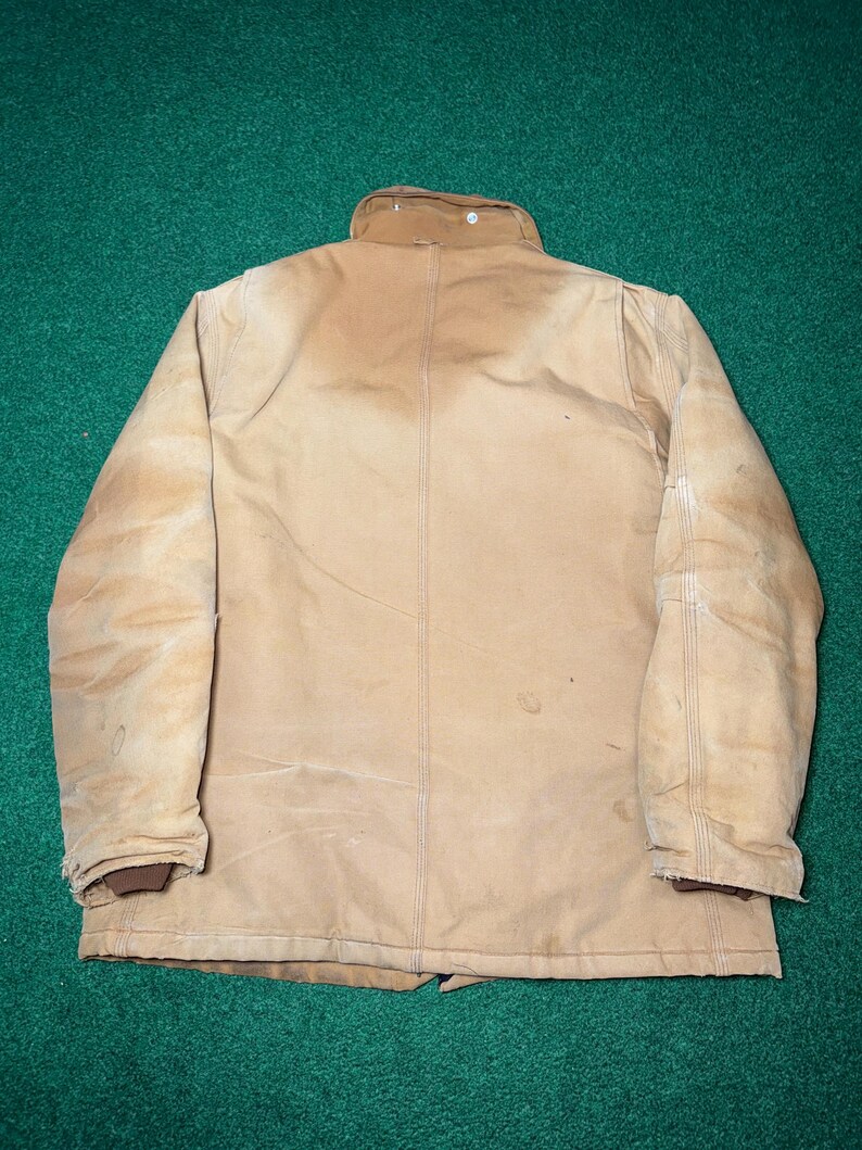 Może przedstawiać: Beżowa kurtka robocza z brązowym kołnierzem i mankietami. Kurtka nosi ślady użytkowania, ze szwami i zapięciem na guziki. Kurtka wykonana jest z wytrzymałego materiału, odpowiedniego do pracy na zewnątrz.