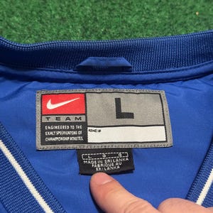 以下が含まれることがあります： 青いナイキジャケットのラベルのクローズアップ。ラベルにはナイキのロゴ、「TEAM」の文字、「ENGINEERED TO THE EXACT SPECIFICATIONS OF CHAMPIONSHIP ATHLETES」のテキストが表示されています。サイズは「L」で、ジャケットはスリランカ製です。