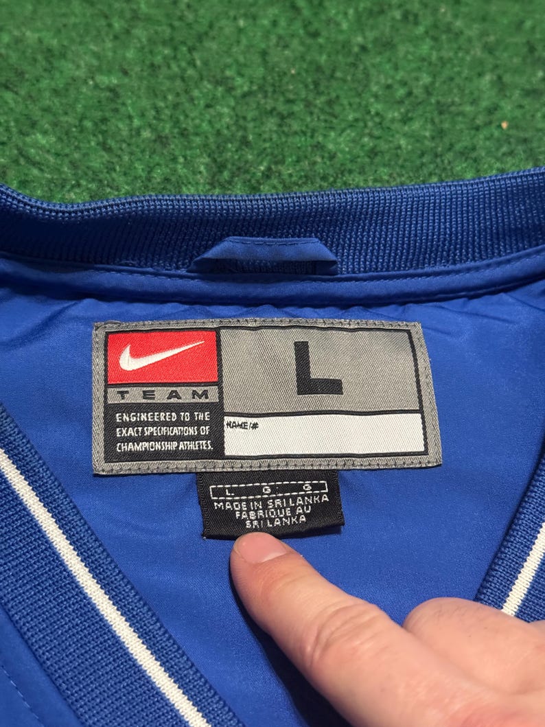 以下が含まれることがあります： 青いナイキジャケットのラベルのクローズアップ。ラベルにはナイキのロゴ、「TEAM」の文字、「ENGINEERED TO THE EXACT SPECIFICATIONS OF CHAMPIONSHIP ATHLETES」のテキストが表示されています。サイズは「L」で、ジャケットはスリランカ製です。