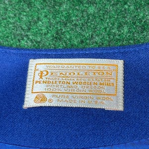 May include: Close-up of blue fabric with a cream-coloured label. The label has gold lettering and reads "PENDLETON WOOLEN MILLS" and "PURE VIRGIN WOOL". It also states "MADE IN U.S.A."