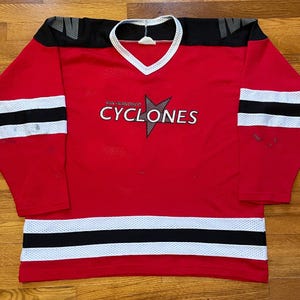 Può includere: Maglia da hockey rossa con strisce bianche e nere. La maglia ha il testo "Saskatoon CYCLONES" sul davanti.