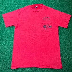 May include: Red short-sleeved t-shirt with a pocket on the front. The pocket has a graphic of a surfer riding a wave. The text "Ron Jon Surf Shop" is printed above the pocket.