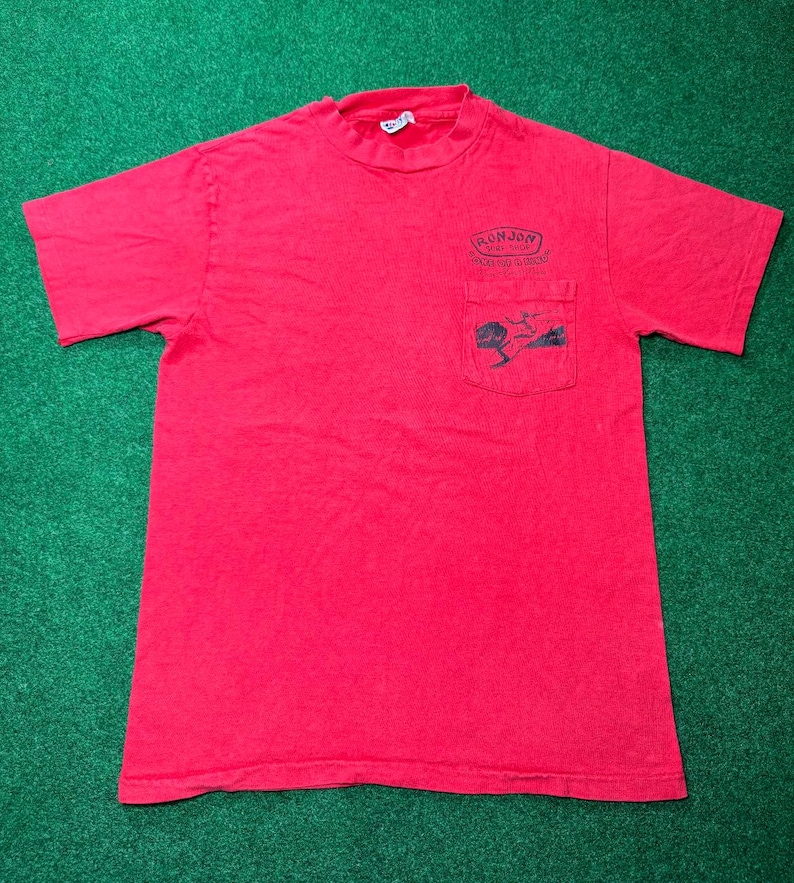 May include: Red short-sleeved t-shirt with a pocket on the front. The pocket has a graphic of a surfer riding a wave. The text "Ron Jon Surf Shop" is printed above the pocket.