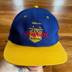 May include: A blue and yellow baseball cap with the embroidered logo "Graybar Racing" and the text "Bussmann" above the logo.