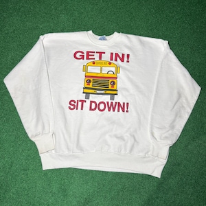 Puede incluir: Sudadera blanca con un diseño de autobús escolar amarillo y negro. Las palabras "GET IN!" y "SIT DOWN!" están escritas en rojo.