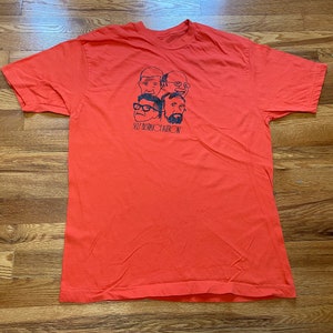 May include: Orange t-shirt with a black and white illustration of five cartoon faces and the text "SELF DESTRUCT BUTTON".