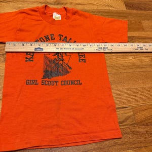 May include: An orange t-shirt with the text "ONE TALL" at the top and "KA EE GIRL SCOUT COUNCIL" at the bottom. A ruler is placed across the shirt. The shirt has short sleeves and a crew neck.
