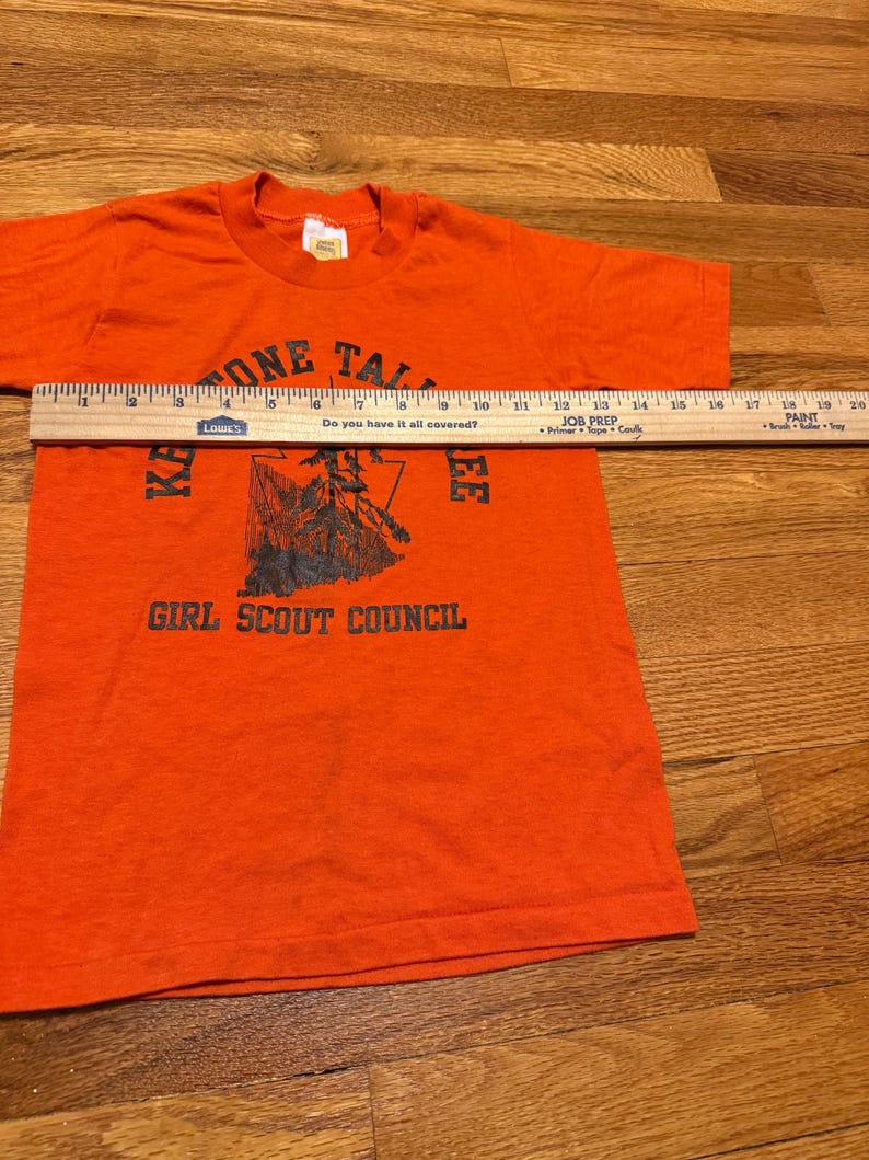 May include: An orange t-shirt with the text "ONE TALL" at the top and "KA EE GIRL SCOUT COUNCIL" at the bottom. A ruler is placed across the shirt. The shirt has short sleeves and a crew neck.