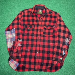 Könnte beinhalten: Rot-schwarz kariertes Flanellhemd mit zwei Brusttaschen. Das Hemd hat ein klassisches Knopfdesign und weist Gebrauchsspuren auf, mit Flicken an den Ärmeln. Das Hemd liegt flach auf einer grünen Oberfläche.