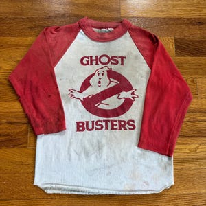 May include: A long-sleeve raglan shirt with a white torso and red sleeves. The front features a red graphic of the Ghostbusters logo with the text GHOST BUSTERS printed in red. The shirt shows signs of wear, including small holes on the right sleeve.