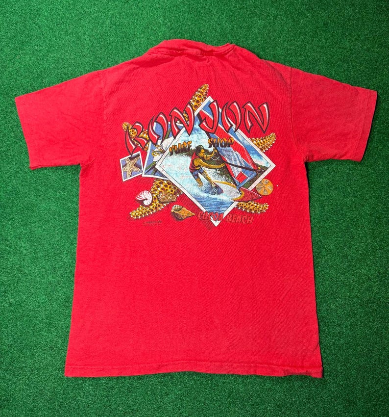 May include: Red t-shirt with a graphic of a person surfing with the text "Ron Jon Surf Shop Cocoa Beach" printed on the back.