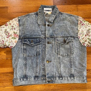 May include: A light blue denim jacket with floral patterned sleeves. The jacket has a button closure and two pockets on the front. The label on the jacket reads "EDG".