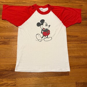 Pu&ograve; includere: T-shirt raglan bianco e rosso con una grafica classica di Topolino. La maglietta ha un corpo bianco con maniche e colletto rossi. L'illustrazione di Topolino &egrave; in nero, rosso e bianco.