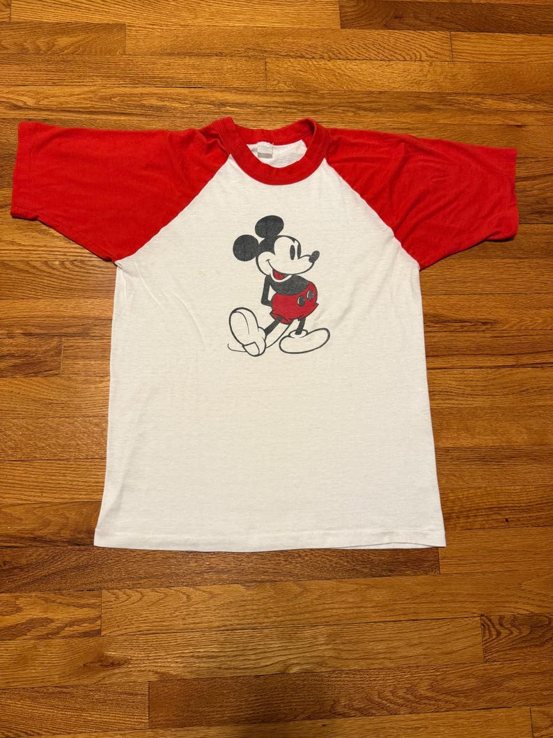 Pu&ograve; includere: T-shirt raglan bianco e rosso con una grafica classica di Topolino. La maglietta ha un corpo bianco con maniche e colletto rossi. L'illustrazione di Topolino &egrave; in nero, rosso e bianco.