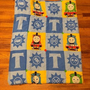 Könnte beinhalten: Eine blaue und gelbe gesteppte Decke mit Thomas der Lokomotive. Die Decke hat ein kariertes Muster mit Bildern von Thomas, einer Eisenbahnstrecke und dem Buchstaben 'T'.