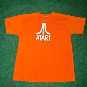 Puede incluir: Una camiseta naranja con un logotipo blanco de Atari que presenta el gráfico clásico del joystick de videojuegos.