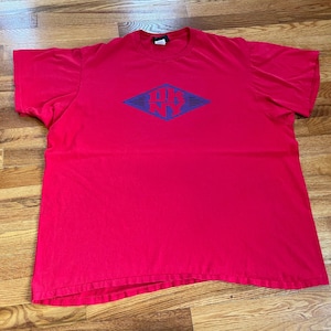 May include: A red t-shirt featuring a blue and white diamond-shaped graphic in the centre. The graphic displays the letters "DK NY". The shirt is made of a soft material and has short sleeves.