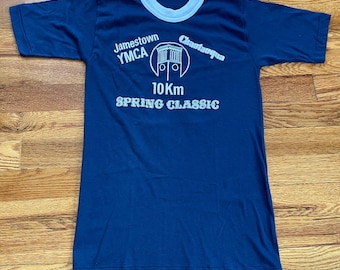 Könnte beinhalten: Ein marineblaues T-Shirt mit einem weißen Ringelkragen. Das Shirt hat einen weißen grafischen Aufdruck mit der Aufschrift "Jamestown YMCA Chautauqua 10Km Spring Classic".