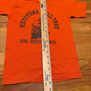 May include: Orange t-shirt with short sleeves. The shirt features the text "KEYSTONE" above a graphic of a tree and the text "GIRL SCOUT COUNCIL". A wooden ruler is placed on the shirt, measuring approximately 22 inches.