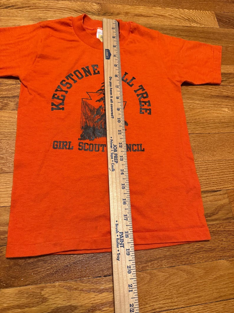 May include: Orange t-shirt with short sleeves. The shirt features the text "KEYSTONE" above a graphic of a tree and the text "GIRL SCOUT COUNCIL". A wooden ruler is placed on the shirt, measuring approximately 22 inches.