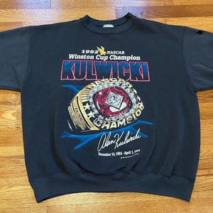 May include: Black sweatshirt featuring a graphic of a gold championship ring and the text "1992 NASCAR Winston Cup Champion Kulwicki" and "Alan Kulwicki" with the dates "December 14, 1954 - April 1, 1993".