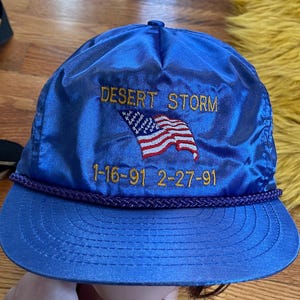 May include: A blue satin baseball cap with the text "Desert Storm" and an American flag embroidered on the front. The dates "1-16-91 2-27-91" are also embroidered on the front.