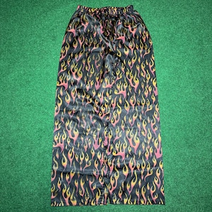 May include: Black skirt with a repeating flame pattern in shades of yellow and pink. The skirt has an elastic waistband and a flowing silhouette. The fabric has a satin sheen, giving it a luxurious appearance. The skirt is laid flat on a green surface.