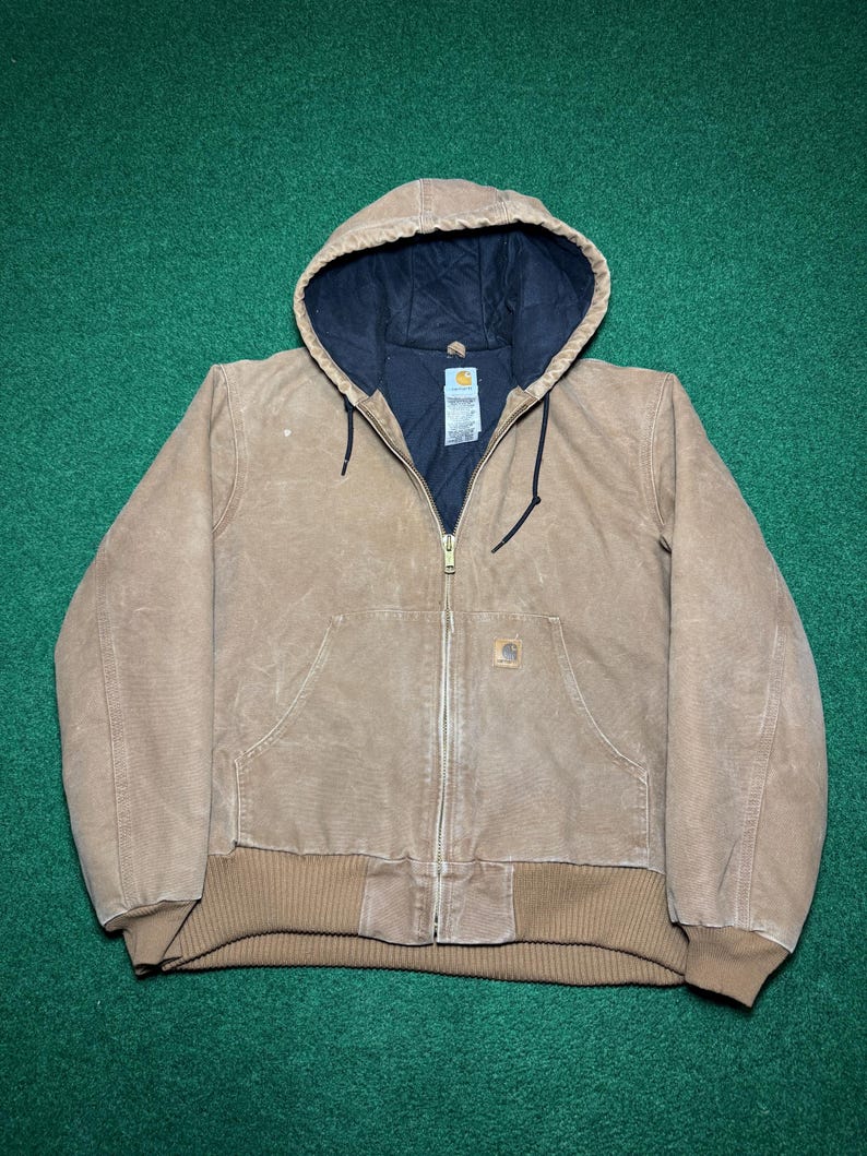 Puede incluir: Chaqueta con capucha marr&oacute;n con forro interior negro. La chaqueta tiene cierre de cremallera, dos bolsillos delanteros y dobladillo y pu&ntilde;os de canal&eacute;. El logotipo de Carhartt es visible en el bolsillo delantero.