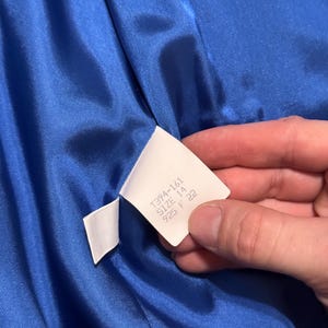 May include: A close-up shot of a bright blue satin fabric. A white label is held against the material, with the text "1394-161 SIZE 14 925 F 22". The fabric is likely part of a piece of clothing.