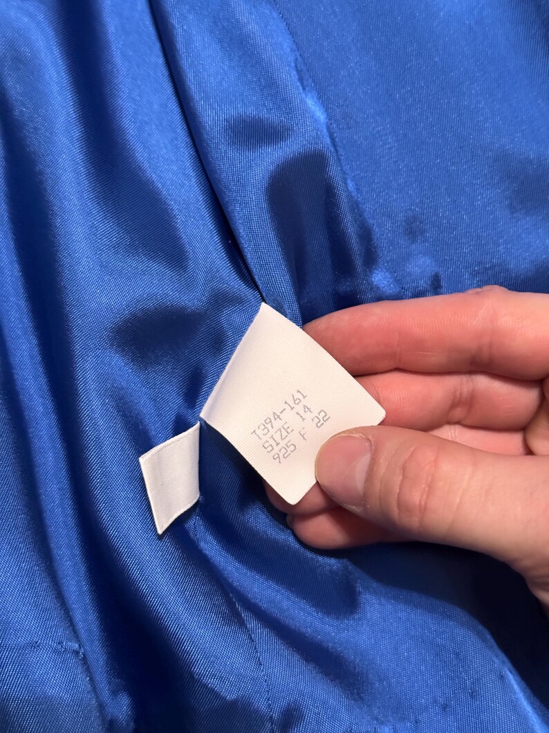 May include: A close-up shot of a bright blue satin fabric. A white label is held against the material, with the text "1394-161 SIZE 14 925 F 22". The fabric is likely part of a piece of clothing.