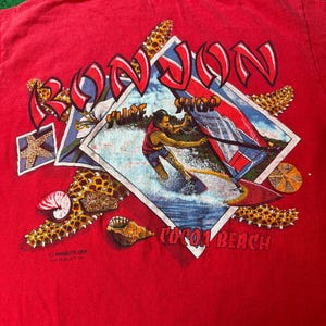 May include: Red t-shirt with a graphic of a person waterskiing in front of a surf shop called "Ron Jon" in Cocoa Beach, Florida. The graphic is surrounded by seashells and starfish.