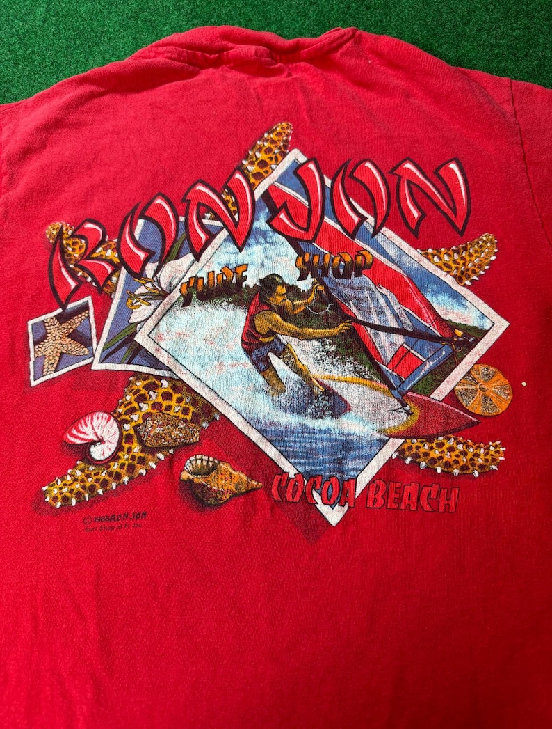 May include: Red t-shirt with a graphic of a person waterskiing in front of a surf shop called "Ron Jon" in Cocoa Beach, Florida. The graphic is surrounded by seashells and starfish.
