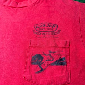 May include: Red t-shirt with a pocket featuring a black and white graphic of a surfer. The shirt has a black and white logo that reads "Ron Jon Surf Shop" and "One of a Kind".