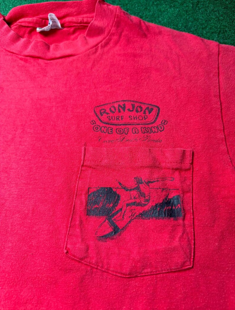 May include: Red t-shirt with a pocket featuring a black and white graphic of a surfer. The shirt has a black and white logo that reads "Ron Jon Surf Shop" and "One of a Kind".