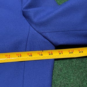 May include: A close-up of a royal blue blazer. The image shows the shoulder and upper sleeve area of the blazer. A yellow measuring tape is placed across the blazer, showing measurements in inches, equivalent to cm.