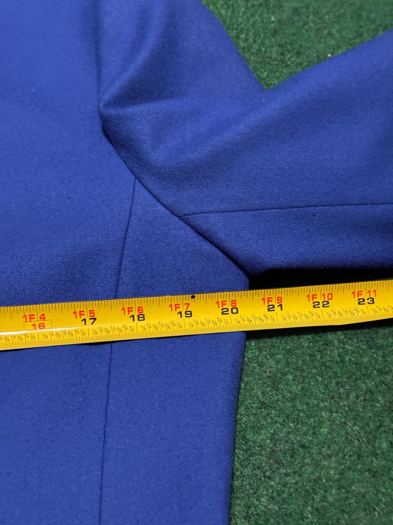 May include: A close-up of a royal blue blazer. The image shows the shoulder and upper sleeve area of the blazer. A yellow measuring tape is placed across the blazer, showing measurements in inches, equivalent to cm.