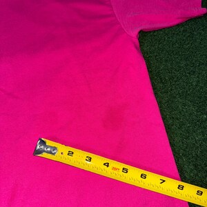 May include: A bright pink t-shirt with a yellow measuring tape placed on top of it. The tape measures 9 inches.