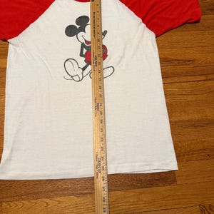 Pu&ograve; includere: T-shirt bianco e rosso a maniche raglan con grafica di Topolino. La maglietta ha un girocollo e un design vintage. Un righello di legno &egrave; posizionato verticalmente accanto alla maglietta, misurando circa 81 cm.