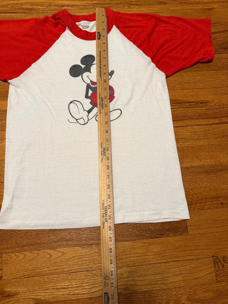 Pu&ograve; includere: T-shirt bianco e rosso a maniche raglan con grafica di Topolino. La maglietta ha un girocollo e un design vintage. Un righello di legno &egrave; posizionato verticalmente accanto alla maglietta, misurando circa 81 cm.