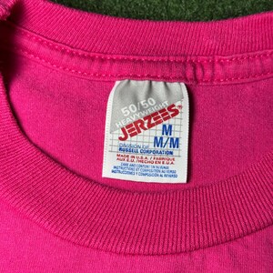 May include: A pink 50/50 heavyweight Jerzees t-shirt with a white tag that reads "50/50 Heavyweight Jerzees" and "M/M Division of Russell Corporation" in black text. The tag also includes care instructions in English and Spanish.