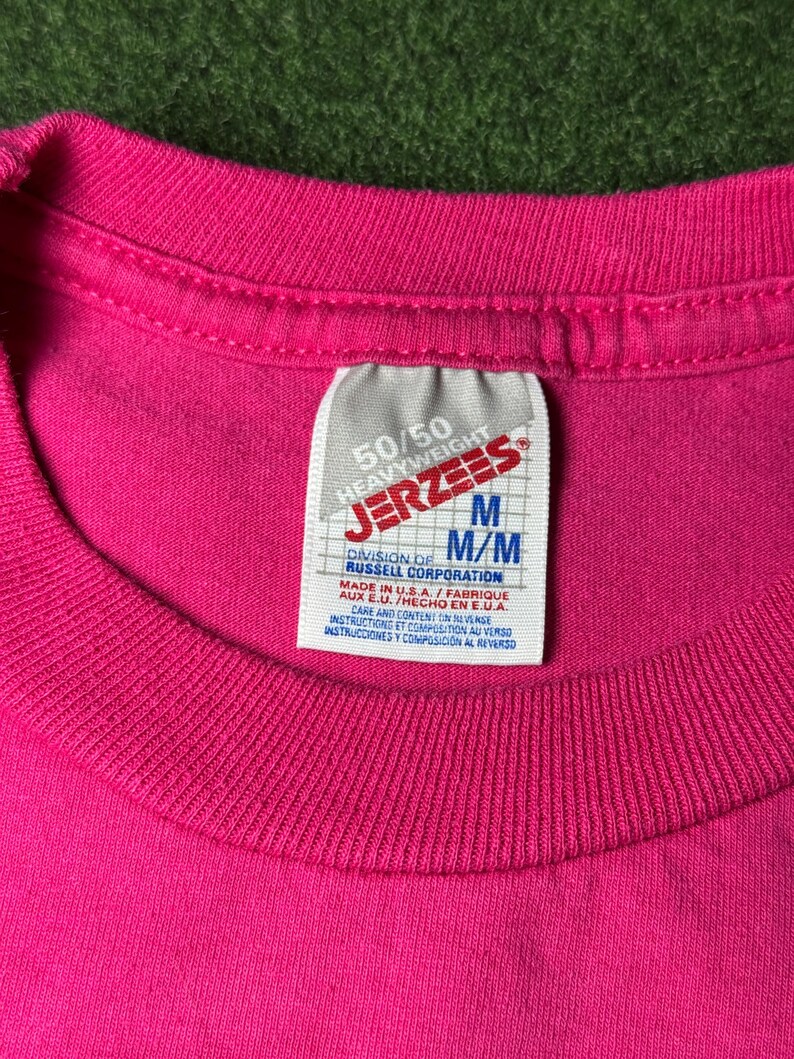 May include: A pink 50/50 heavyweight Jerzees t-shirt with a white tag that reads "50/50 Heavyweight Jerzees" and "M/M Division of Russell Corporation" in black text. The tag also includes care instructions in English and Spanish.