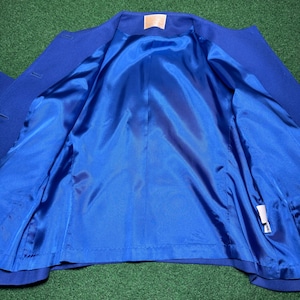 May include: A royal blue blazer with a bright blue satin lining. The inside of the jacket is visible, showing the smooth, shiny fabric. The blazer is displayed on a green surface.