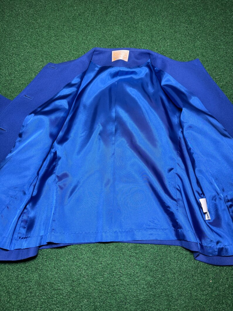 May include: A royal blue blazer with a bright blue satin lining. The inside of the jacket is visible, showing the smooth, shiny fabric. The blazer is displayed on a green surface.