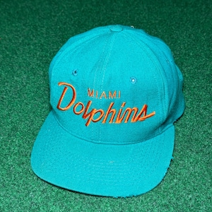 Op de afbeelding: Een groenblauwe honkbalpet met de tekst MIAMI Dolphins in oranje geborduurd op de voorzijde. De pet heeft een gestructureerde kroon met ventilatiegaatjes en een platte klep. Het ontwerp is consistent met professionele sportmerchandise.