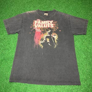 May include: Dark gray t-shirt featuring the text "The Family Values Tour 2007" with a graphic of a person. The design includes red, pink, and yellow accents. The shirt is made of a soft material.
