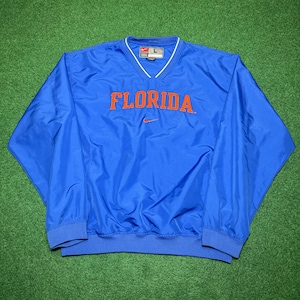 以下が含まれることがあります： 胸にオレンジ色のブロック体で「FLORIDA」の文字が入った青いナイキのウィンドブレーカー。ウィンドブレーカーは、白いトリムと小さなナイキのロゴが付いたVネックです。袖と裾は伸縮性があります。