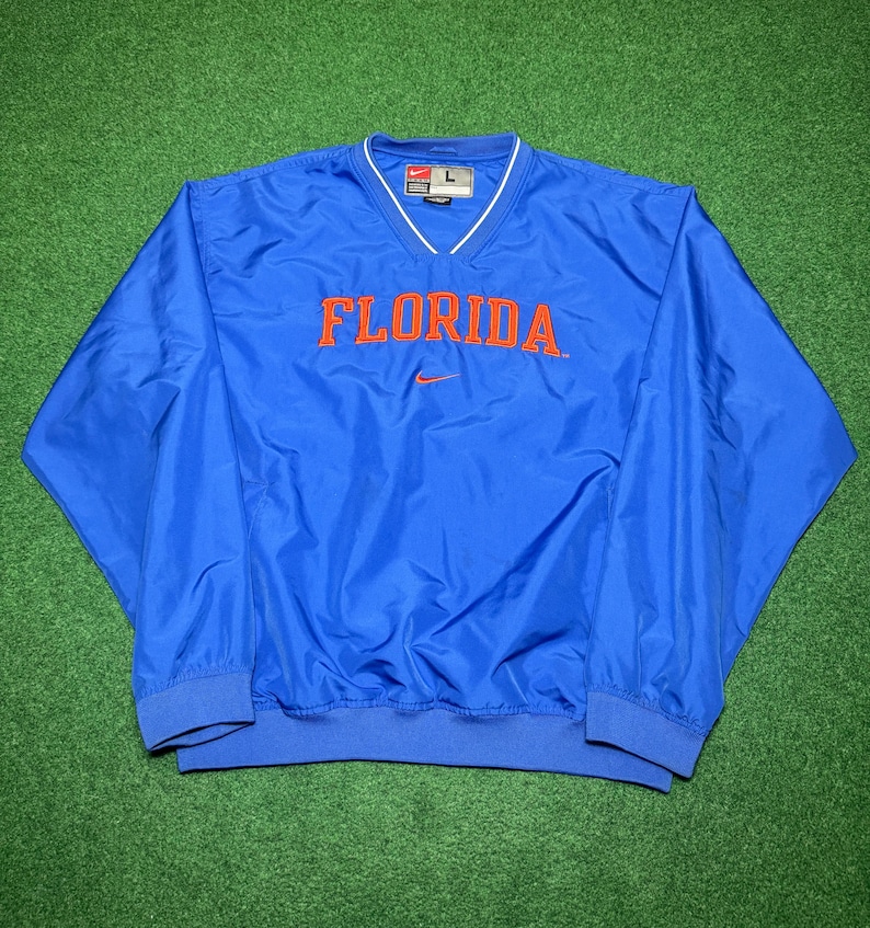 以下が含まれることがあります： 胸にオレンジ色のブロック体で「FLORIDA」の文字が入った青いナイキのウィンドブレーカー。ウィンドブレーカーは、白いトリムと小さなナイキのロゴが付いたVネックです。袖と裾は伸縮性があります。
