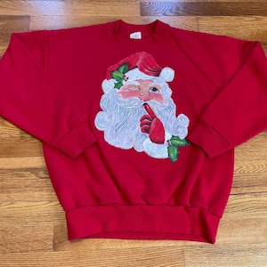 May include: Red Christmas sweatshirt featuring a painted image of Santa Claus with a white beard and a red hat. Santa is winking and holding his finger to his lips.